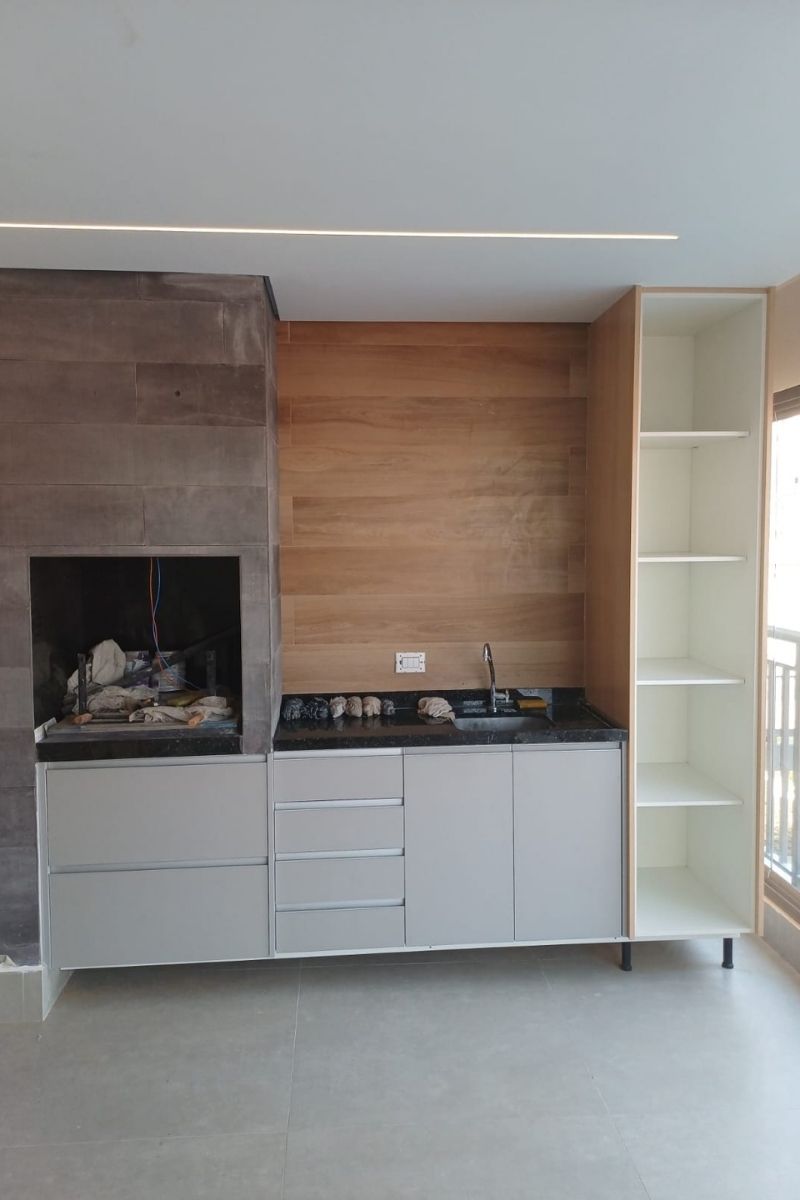 Varanda gourmet planejada em apartamento com churrasqueira, armários brancos sob medida e revestimento de parede em madeira.