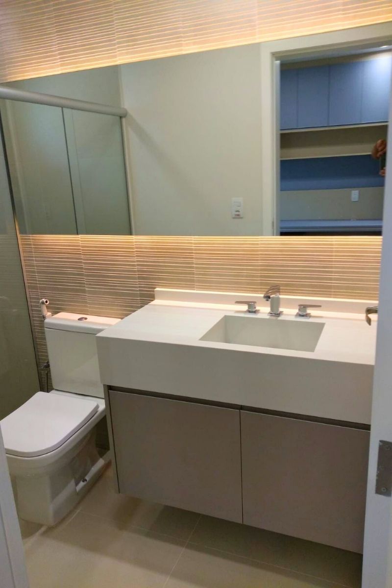 Lavabo de alto padrão com bancada branca extensa, espelho horizontal iluminado por LED e revestimento 3D na parede.