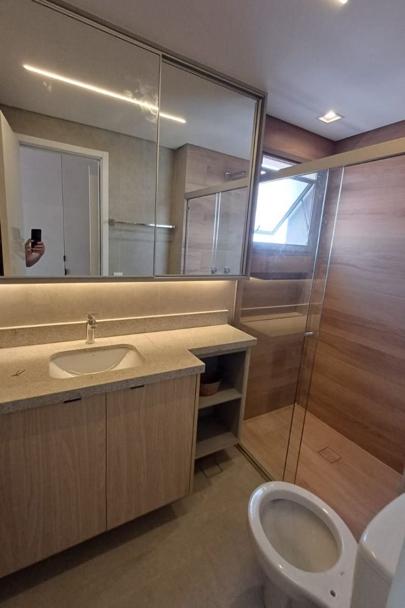 Banheiro planejado completo com gabinete amadeirado, bancada de pedra clara e grande espelho com iluminação LED inferior.