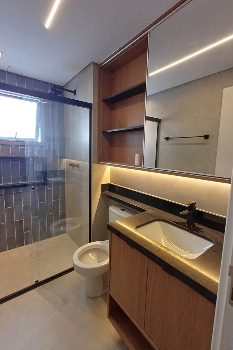 Banheiro planejado moderno com nichos de madeira sobre a caixa acoplada, espelho com iluminação LED e revestimento escuro.