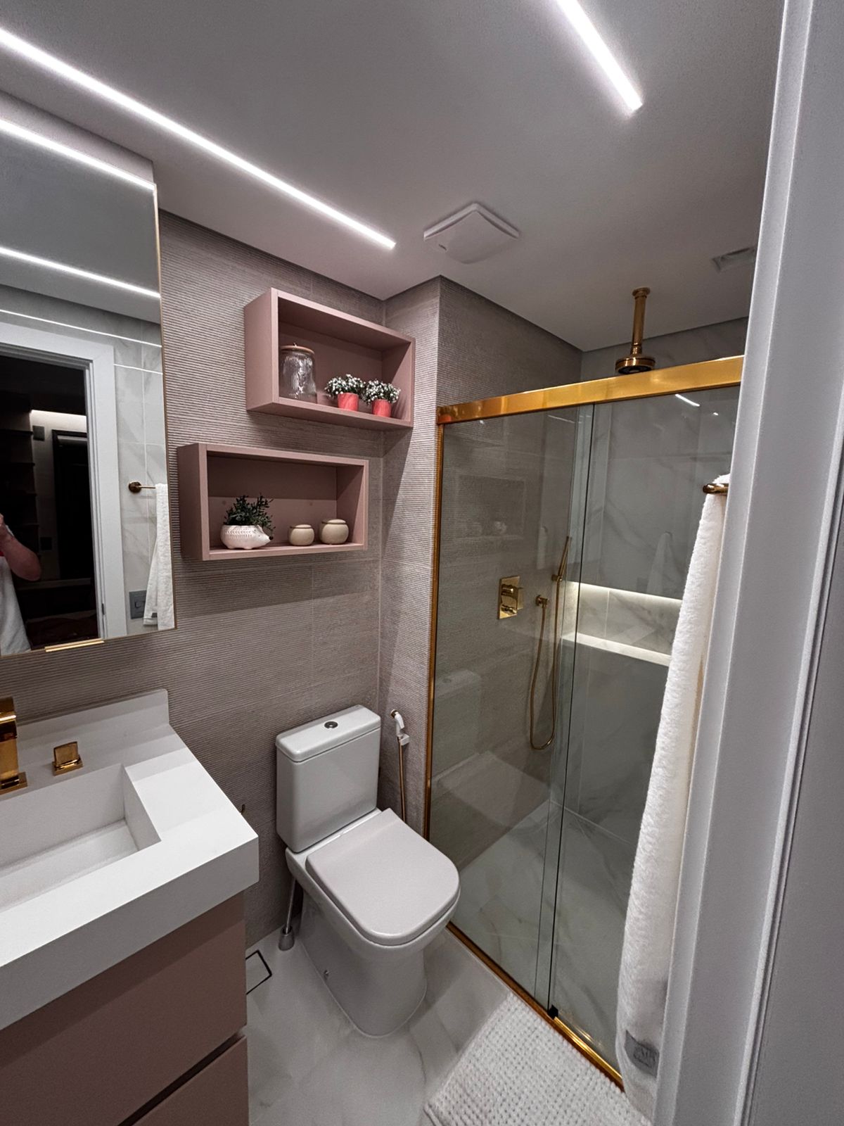 Banheiro planejado moderno com nichos de madeira sobre a caixa acoplada, espelho com iluminação LED e revestimento escuro.