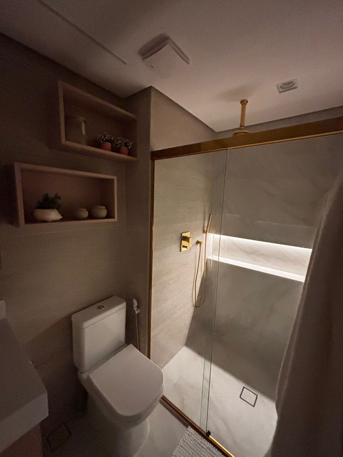 Banheiro planejado moderno com nichos de madeira sobre a caixa acoplada, espelho com iluminação LED e revestimento escuro.