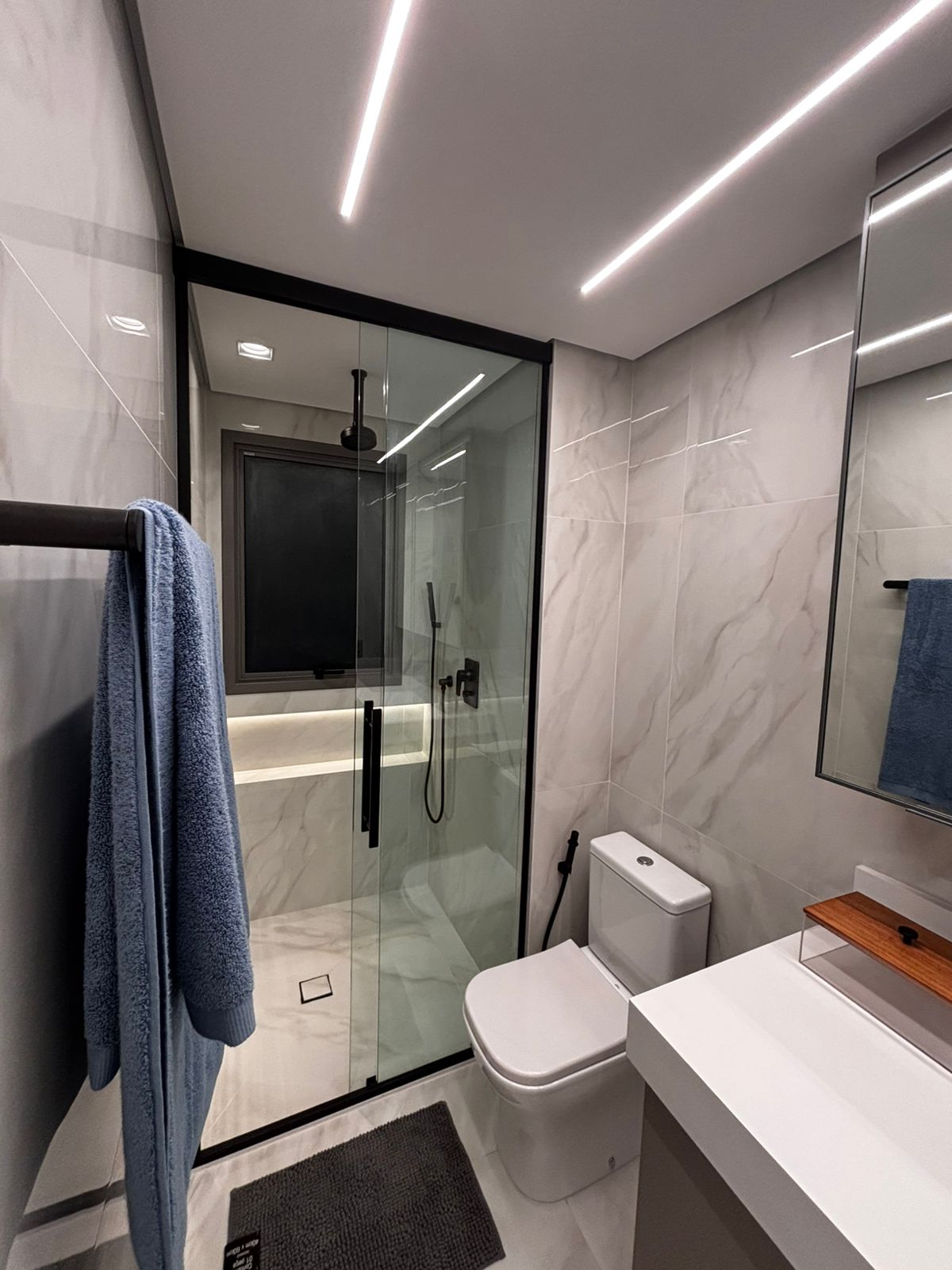 Banheiro planejado moderno com nichos de madeira sobre a caixa acoplada, espelho com iluminação LED e revestimento escuro.