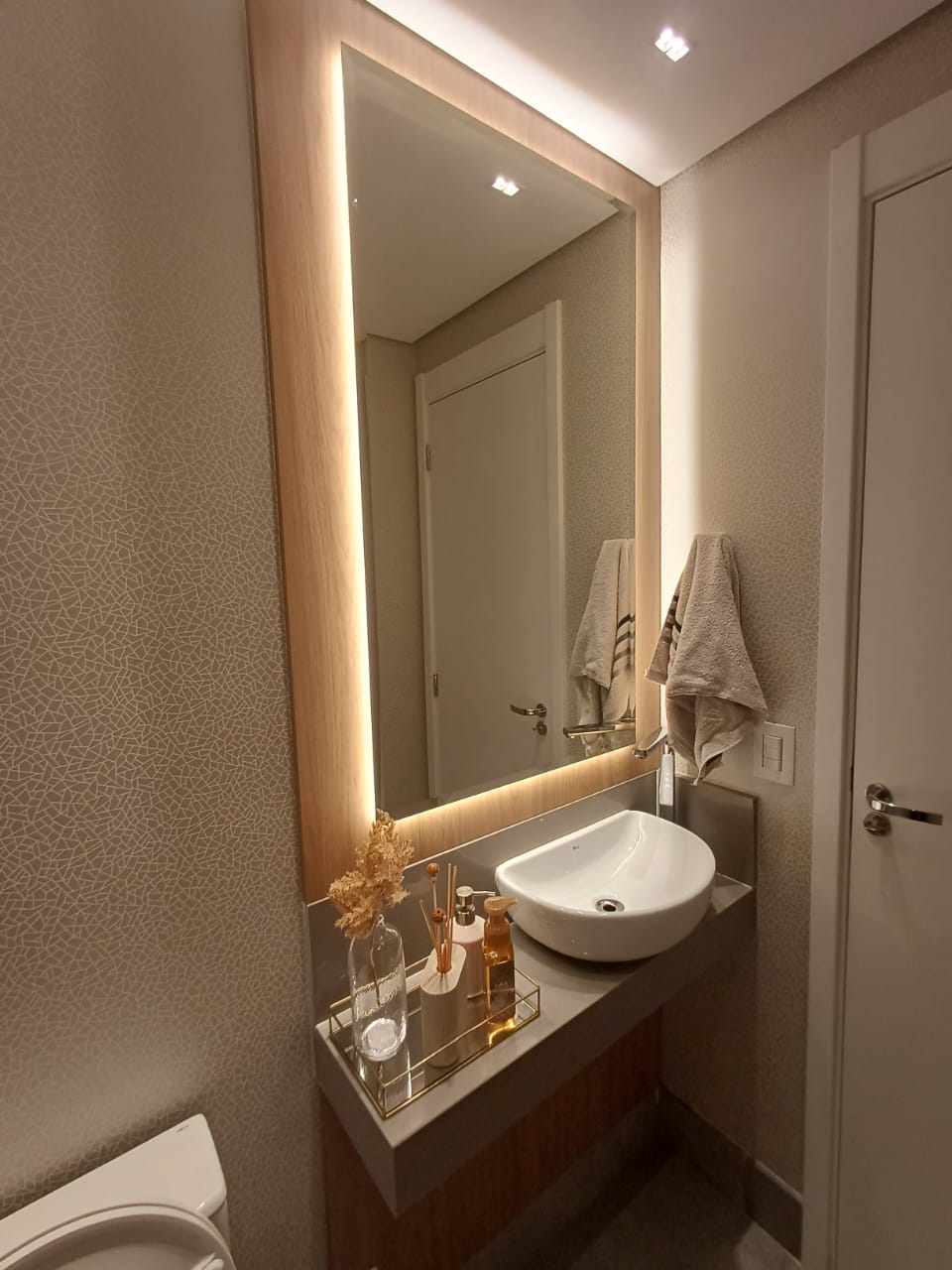 Lavabo de alto padrão com bancada branca extensa, espelho horizontal iluminado por LED e revestimento 3D na parede.