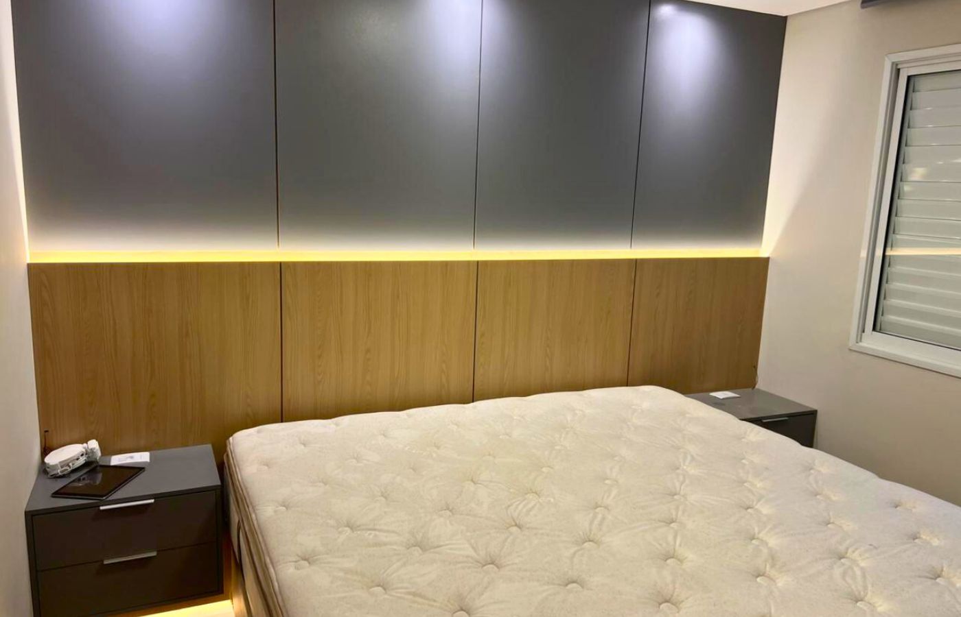 Quarto de casal planejado com cabeceira em madeira ripada, iluminação LED indireta (fita de LED) e armários superiores cinza.