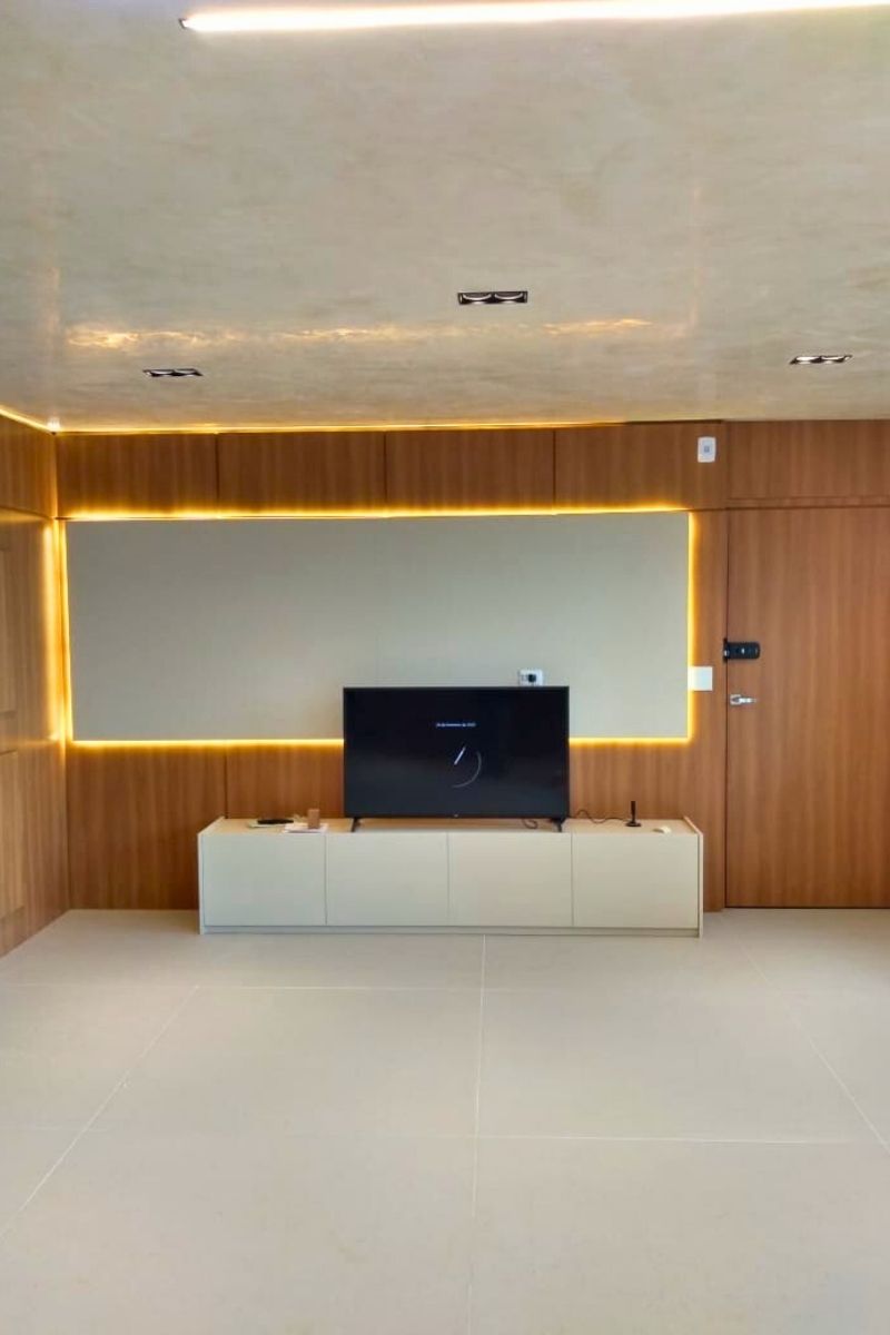Painel de TV com iluminação indireta (backlight) em fita de LED, rack branco e parede revestida em madeira.